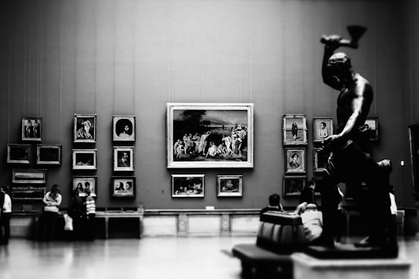A large gallery room featuring several framed artworks mounted on the walls, including portraits and scenes. A prominent statue of a muscular figure with raised arm is positioned in the foreground on the right. Several visitors, some standing and others seated, are admiring the art pieces.