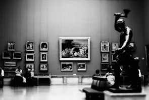 A large gallery room featuring several framed artworks mounted on the walls, including portraits and scenes. A prominent statue of a muscular figure with raised arm is positioned in the foreground on the right. Several visitors, some standing and others seated, are admiring the art pieces.