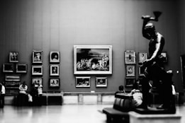A large gallery room featuring several framed artworks mounted on the walls, including portraits and scenes. A prominent statue of a muscular figure with raised arm is positioned in the foreground on the right. Several visitors, some standing and others seated, are admiring the art pieces.