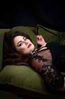 A woman with striking makeup and long, dark hair is lying on a green velvet sofa. She is wearing a lace dress that partially reveals her shoulder, and there is a spiked choker around her neck. The setting is dimly lit, creating a dramatic and intimate atmosphere.