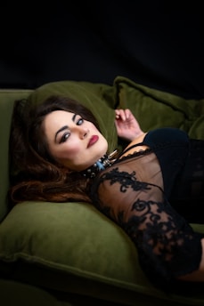 A woman with striking makeup and long, dark hair is lying on a green velvet sofa. She is wearing a lace dress that partially reveals her shoulder, and there is a spiked choker around her neck. The setting is dimly lit, creating a dramatic and intimate atmosphere.