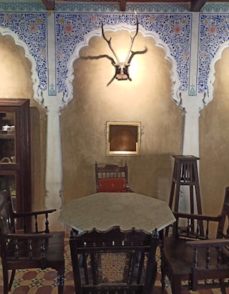 A traditional room featuring ornate archways with intricate blue and white floral patterns. There is a mounted antelope head with antlers on the wall, and a small square niche below it. Furniture in the room includes an octagonal table surrounded by wooden chairs, and a tall wooden stand. The floor has a patterned tile design.