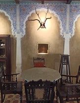 A traditional room featuring ornate archways with intricate blue and white floral patterns. There is a mounted antelope head with antlers on the wall, and a small square niche below it. Furniture in the room includes an octagonal table surrounded by wooden chairs, and a tall wooden stand. The floor has a patterned tile design.
