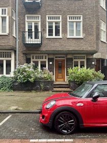 A three-story brick building features large windows with white frames and black metal railings on the second floor. A small garden with hydrangeas is in front of the entrance, leading to a wooden door with a glass panel. A red car is parked on the street, with wet paving stones suggesting recent rain.