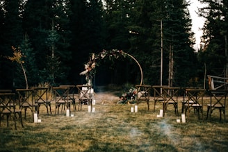 An outdoor wedding setup with wooden chairs arranged in rows on a grassy area. In the background, there is a circular floral arch surrounded by trees. Soft lighting is provided by candles placed on the ground.