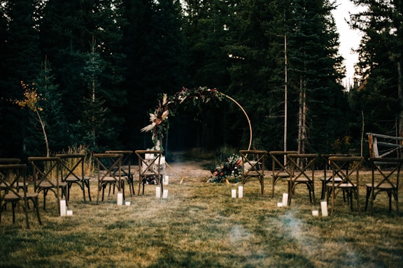 An outdoor wedding setup with wooden chairs arranged in rows on a grassy area. In the background, there is a circular floral arch surrounded by trees. Soft lighting is provided by candles placed on the ground.