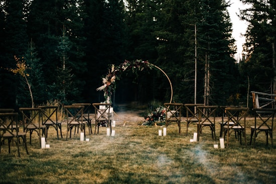 An outdoor wedding setup with wooden chairs arranged in rows on a grassy area. In the background, there is a circular floral arch surrounded by trees. Soft lighting is provided by candles placed on the ground.