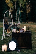 A dark and somewhat eerie outdoor setup is depicted, featuring a small table adorned with various objects. Items on the table include a human skull, a bouquet of flowers, several bottles of alcohol, candles, and a small birdcage. In front of the table, a decorative mirror with a black frame rests on the grass along with several large lit candles. In the background, a tall white framed mirror stands on the grass, reflecting the surrounding trees and foliage, which hint at early evening or dusk.