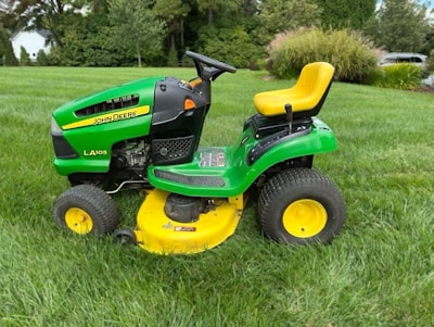 A bright green and yellow ride-on lawn mower sits on a well-manicured lawn, surrounded by lush greenery and trees in the background.