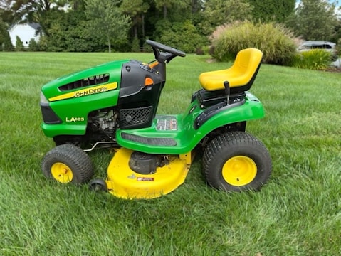 A bright green and yellow ride-on lawn mower sits on a well-manicured lawn, surrounded by lush greenery and trees in the background.