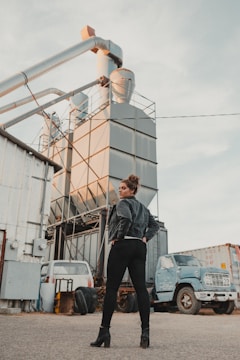 A person stands confidently with a hand on their hip in front of industrial machinery. The background contains metal structures and large pipes, along with two parked, worn-out vehicles.