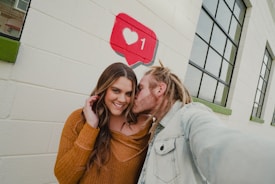 A person with long brown hair, wearing a brown sweater, smiles while another person with dreadlocks and a denim jacket kisses them on the cheek. They stand against a painted exterior brick wall with large windows, which features a red and white social media-like icon with a heart and number 1.