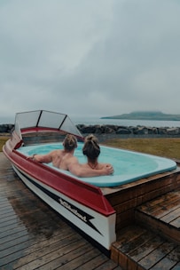 A unique hot tub designed in the shape of a boat, with two people relaxing inside. The setting appears to be by the seaside, with a misty and overcast sky in the background. The deck surrounding the hot tub is made of wooden planks.
