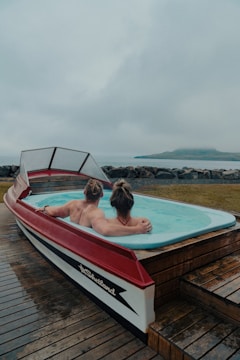 A unique hot tub designed in the shape of a boat, with two people relaxing inside. The setting appears to be by the seaside, with a misty and overcast sky in the background. The deck surrounding the hot tub is made of wooden planks.