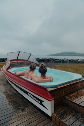 A unique hot tub designed in the shape of a boat, with two people relaxing inside. The setting appears to be by the seaside, with a misty and overcast sky in the background. The deck surrounding the hot tub is made of wooden planks.