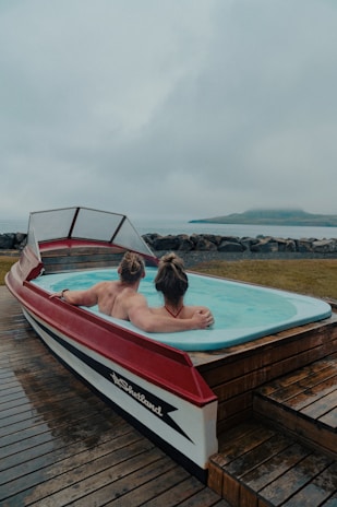 A unique hot tub designed in the shape of a boat, with two people relaxing inside. The setting appears to be by the seaside, with a misty and overcast sky in the background. The deck surrounding the hot tub is made of wooden planks.