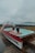 A unique hot tub designed in the shape of a boat, with two people relaxing inside. The setting appears to be by the seaside, with a misty and overcast sky in the background. The deck surrounding the hot tub is made of wooden planks.