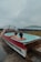 A unique hot tub designed in the shape of a boat, with two people relaxing inside. The setting appears to be by the seaside, with a misty and overcast sky in the background. The deck surrounding the hot tub is made of wooden planks.