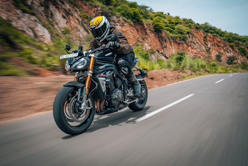 A motorcyclist wearing protective gear and a helmet rides a sports motorcycle on a scenic road. The road is bordered by rocky terrain and green vegetation, indicating a natural and possibly mountainous area. The motorcycle is moving at high speed, which is captured by the motion blur in the image.