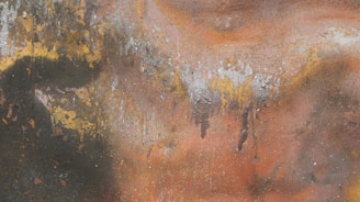 A textured surface featuring layers of rust and peeling paint. The colors blend together in organic patterns, exhibiting various shades influenced by corrosion and weathering. The surface displays patches of dark green, yellow, orange, and gray with irregular spots and streaks.