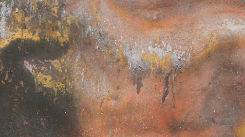 A textured surface featuring layers of rust and peeling paint. The colors blend together in organic patterns, exhibiting various shades influenced by corrosion and weathering. The surface displays patches of dark green, yellow, orange, and gray with irregular spots and streaks.
