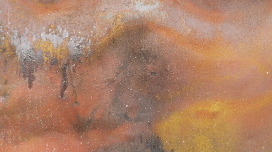 A textured surface exhibits a rich array of organic patterns with an earthy palette. The surface seems to have layers of rust and corrosion, creating a rugged and uneven aesthetic. Shades of orange, brown, yellow, and gray dominate, with specks of white scattered throughout, adding depth and complexity.