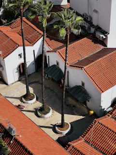 An aerial view of a courtyard with several tall palm trees growing in circular planters. The courtyard is surrounded by buildings with pitched roofs covered in red tiles. The architecture features white walls, a tiled pathway, and traditional design elements. Some small objects are placed around the courtyard, possibly outdoor furniture or decorations.