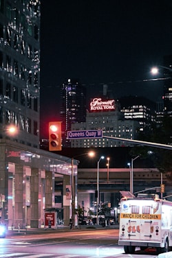 City street scene at night featuring illuminated signs, including a prominent hotel sign in red that reads 'Fairmont Royal York'. A street intersection can be seen with a traffic light turned red, and a street sign labeled 'Queens Quay W'. An illuminated vehicle in the foreground has a 'Watch for Children' sign on the back. Modern buildings with reflective windows and additional signage are visible in the background.