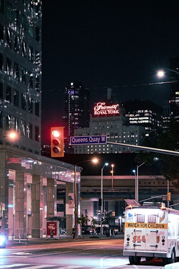 City street scene at night featuring illuminated signs, including a prominent hotel sign in red that reads 'Fairmont Royal York'. A street intersection can be seen with a traffic light turned red, and a street sign labeled 'Queens Quay W'. An illuminated vehicle in the foreground has a 'Watch for Children' sign on the back. Modern buildings with reflective windows and additional signage are visible in the background.
