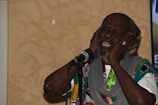 An elderly man with a beard is expressing deep emotion through his singing. He has his eyes closed and is holding his head with both hands near a microphone. He is wearing a multicolored patterned garment and a gray scarf.