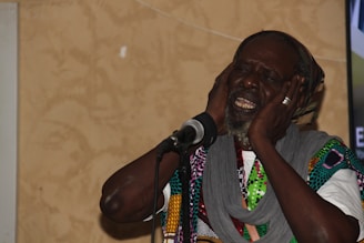 An elderly man with a beard is expressing deep emotion through his singing. He has his eyes closed and is holding his head with both hands near a microphone. He is wearing a multicolored patterned garment and a gray scarf.