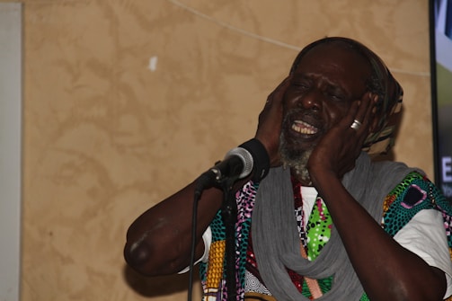 An elderly man with a beard is expressing deep emotion through his singing. He has his eyes closed and is holding his head with both hands near a microphone. He is wearing a multicolored patterned garment and a gray scarf.