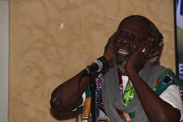 An elderly man with a beard is expressing deep emotion through his singing. He has his eyes closed and is holding his head with both hands near a microphone. He is wearing a multicolored patterned garment and a gray scarf.
