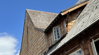 A close-up view of a wooden building featuring steeply sloped roofs covered with shingles. The building has multiple windows set at angles due to the varying roof heights. The sky is clear and blue, suggesting calm weather.