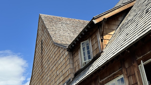 A close-up view of a wooden building featuring steeply sloped roofs covered with shingles. The building has multiple windows set at angles due to the varying roof heights. The sky is clear and blue, suggesting calm weather.