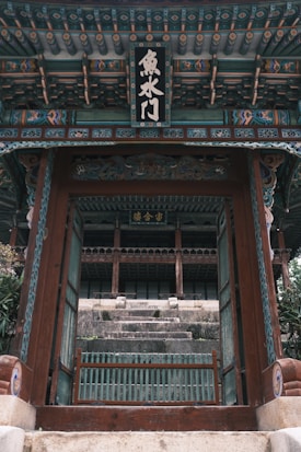 A traditional wooden gate with intricate patterns and calligraphy leads to an ancient temple with stone steps. The temple's architecture features ornate details in muted colors, reflecting cultural heritage.