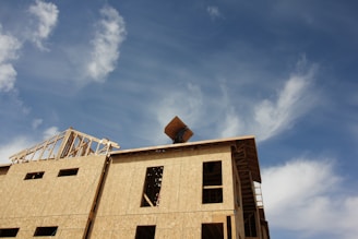 A partially constructed building with wooden frames and bare walls under a clear blue sky with scattered clouds. A construction crane is visible in the background, lifting materials.