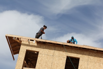 Two individuals are working on the roof of an unfinished wooden structure. One person is standing and appears to be handling something with a cable, while the other is sitting, wearing a blue outfit with a hood. The sky is partly cloudy, adding a sense of openness to the scene.