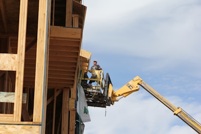 A construction worker stands on an elevated platform beside a wooden structure under construction. The worker wears protective gear and operates equipment attached to a yellow crane-like machine. The backdrop is a clear, slightly cloudy sky.
