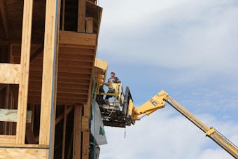 A construction worker stands on an elevated platform beside a wooden structure under construction. The worker wears protective gear and operates equipment attached to a yellow crane-like machine. The backdrop is a clear, slightly cloudy sky.