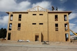 A multi-story building under construction with wooden boards covering the exterior. Two workers are visible on the roof, and various construction materials are scattered around the site. The frame of the roof is partially completed against a backdrop of blue sky and clouds.