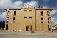 A multi-story building under construction with wooden boards covering the exterior. Two workers are visible on the roof, and various construction materials are scattered around the site. The frame of the roof is partially completed against a backdrop of blue sky and clouds.
