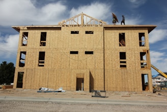 A multi-story building under construction with wooden boards covering the exterior. Two workers are visible on the roof, and various construction materials are scattered around the site. The frame of the roof is partially completed against a backdrop of blue sky and clouds.