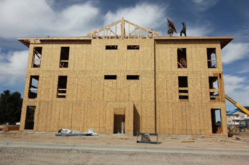 A multi-story building under construction with wooden boards covering the exterior. Two workers are visible on the roof, and various construction materials are scattered around the site. The frame of the roof is partially completed against a backdrop of blue sky and clouds.