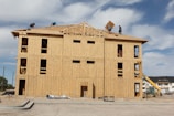 A multi-story wooden building under construction is shown with several workers on the roof working on the structure. The building appears to be framed with large sheets of plywood, and a construction vehicle is present on the right side. The sky is clear with a few clouds.