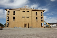 A partially constructed building with wooden walls and exposed frameworks stands on a construction site. Two workers are visible on the roof. The ground is covered with gravel, and construction equipment, including a telescopic handler, is nearby. The background shows blue sky with scattered clouds.