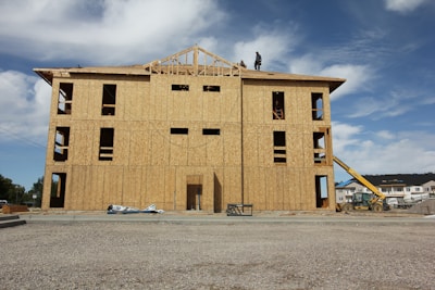 A partially constructed building with wooden walls and exposed frameworks stands on a construction site. Two workers are visible on the roof. The ground is covered with gravel, and construction equipment, including a telescopic handler, is nearby. The background shows blue sky with scattered clouds.
