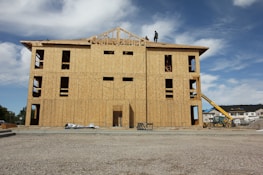 A partially constructed building with wooden walls and exposed frameworks stands on a construction site. Two workers are visible on the roof. The ground is covered with gravel, and construction equipment, including a telescopic handler, is nearby. The background shows blue sky with scattered clouds.