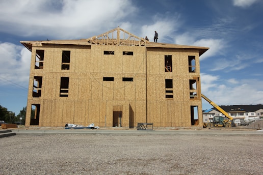 A partially constructed building with wooden walls and exposed frameworks stands on a construction site. Two workers are visible on the roof. The ground is covered with gravel, and construction equipment, including a telescopic handler, is nearby. The background shows blue sky with scattered clouds.