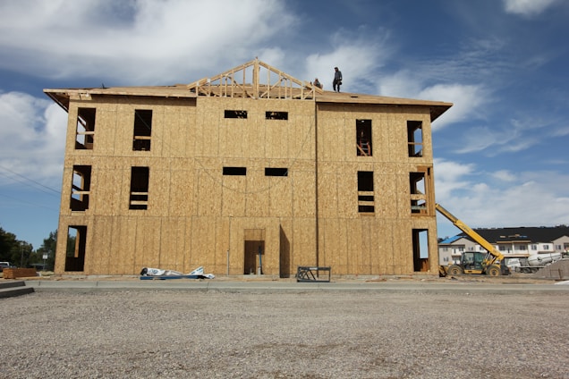 A partially constructed building with wooden walls and exposed frameworks stands on a construction site. Two workers are visible on the roof. The ground is covered with gravel, and construction equipment, including a telescopic handler, is nearby. The background shows blue sky with scattered clouds.